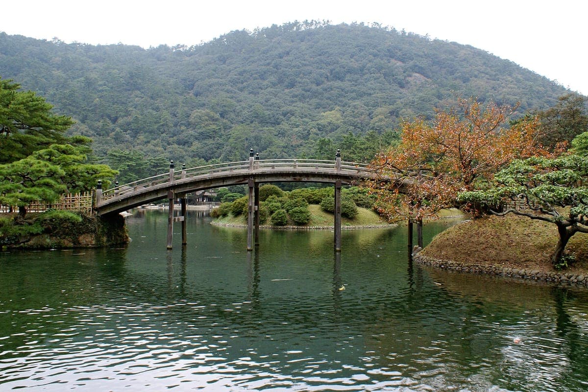 Eine gewölbte Holzbrücke überquert einen noch grünen Teich im Ritsurin-Garten mit geschnitzten Kiefern, einem rotblättrigen Baum und einem bewaldeten Berg dahinter