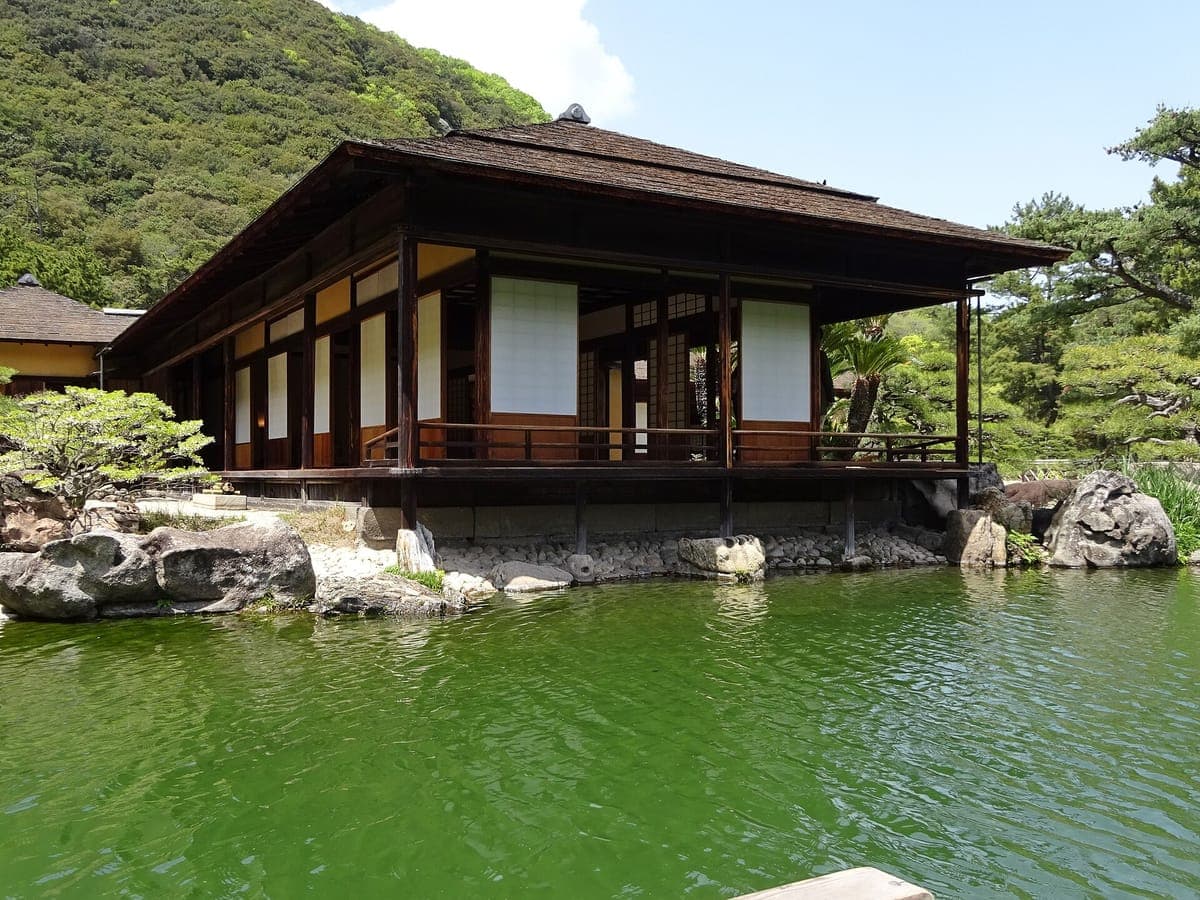 Das hölzerne Kikugetsu-tei Teehaus mit tiefem Ziegeldach und offener Veranda über einem grünen Teich mit Felsen und beschnittenen Kiefern am Ufer