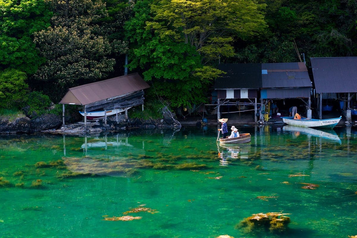 Een kleine boot met ronde kuip op helder smaragdgroen water in een rotsachtige baai bij Yajima-Kyōjima op Sado, met daarachter houten bootschuren tussen groene bomen