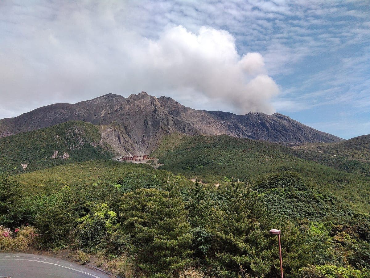 Ang tulis-tulis na kulay-abo na mga taluktok ng Sakurajima na tumataas sa ibabaw ng berdeng kagubatan na dalisdis, na may makapal na puting usok na umaagos mula sa tuktok