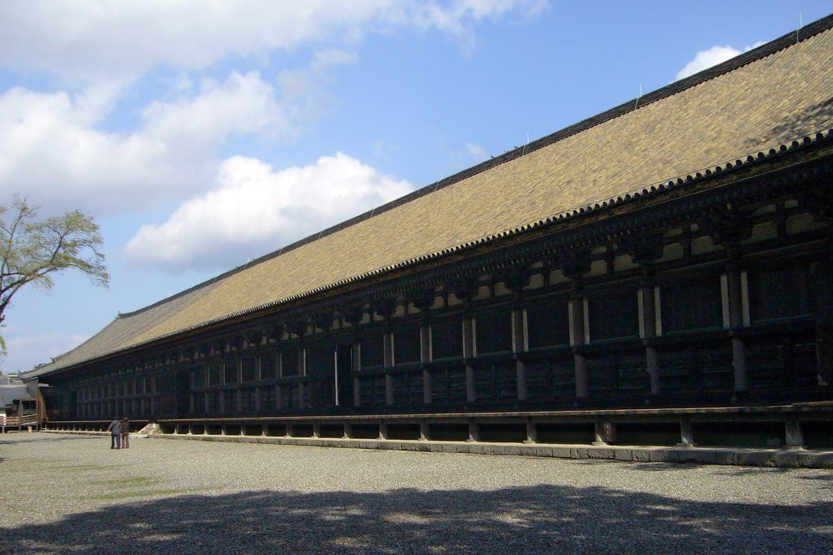 El larguísimo pabellón principal de madera de Sanjūsangen-dō con un techo bajo de tejas que se extiende a lo lejos a través de un patio de grava bajo un cielo azul