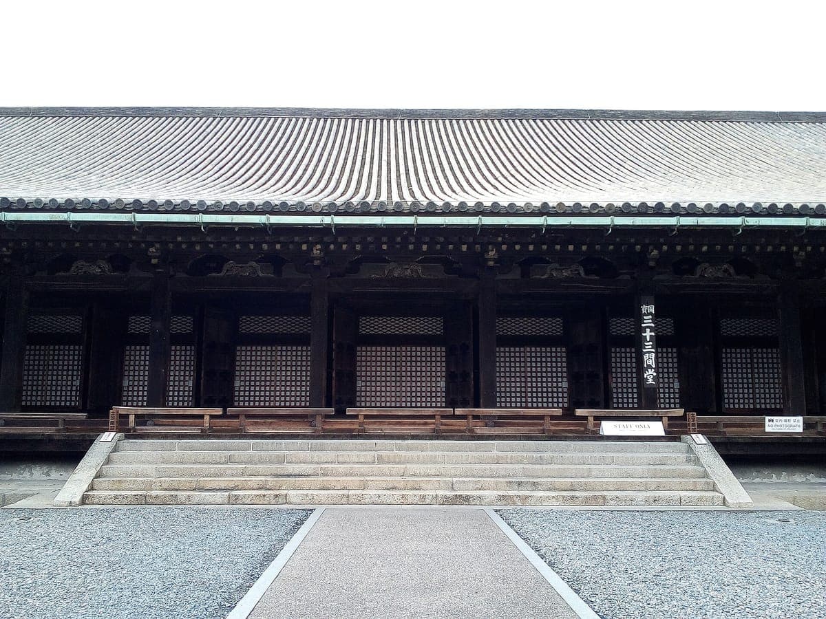 El frente del Sanjūsangen-dō pabellón principal con puertas de celosía de madera oscura, un largo techo de tejas, escalones de piedra que conducen hacia arriba y un letrero vertical que dice Sanjūsangen-dō al lado de un aviso de no tomar fotografías.