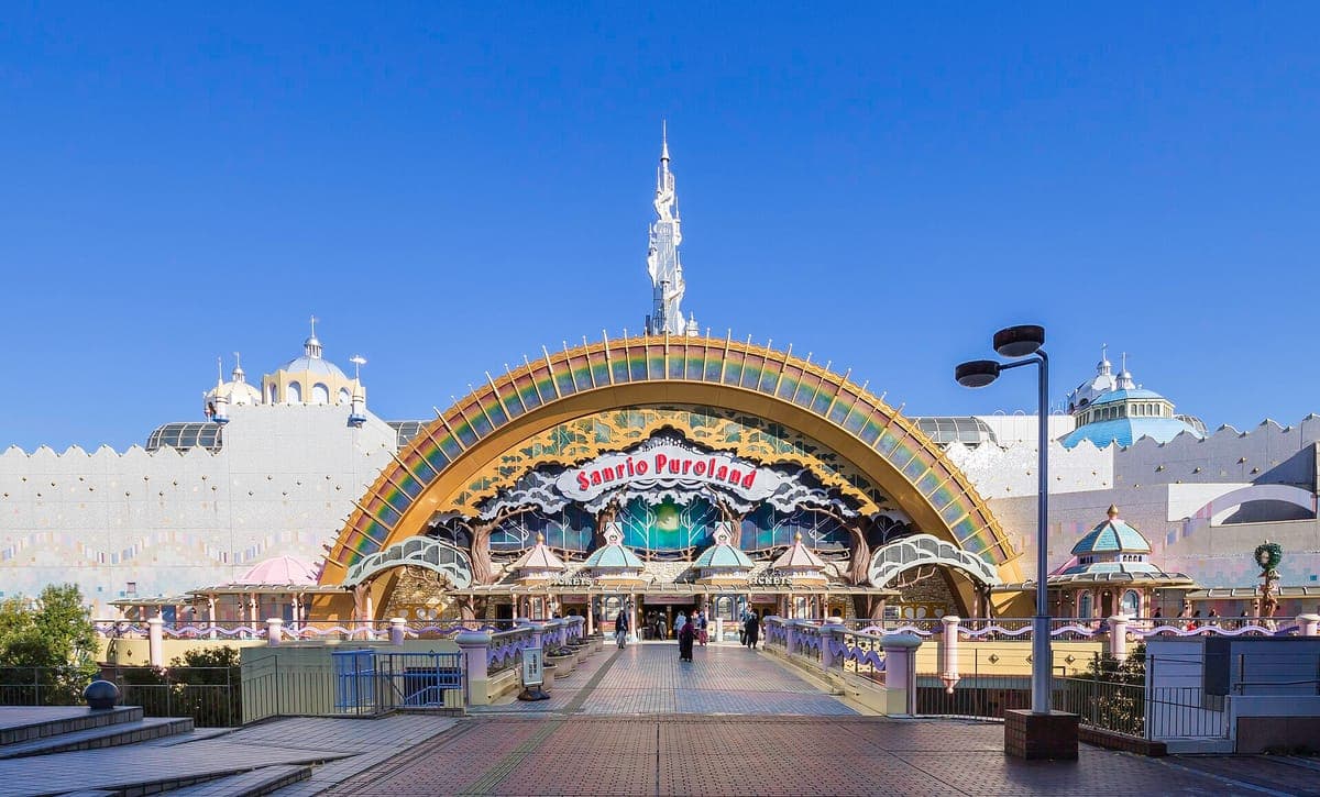 Die regenbogengewölbte Eingangsfassade von Sanrio Puroland mit einem hohen weißen Turm, Kuppeln und einem breiten gepflasterten Zugang unter blauem Himmel