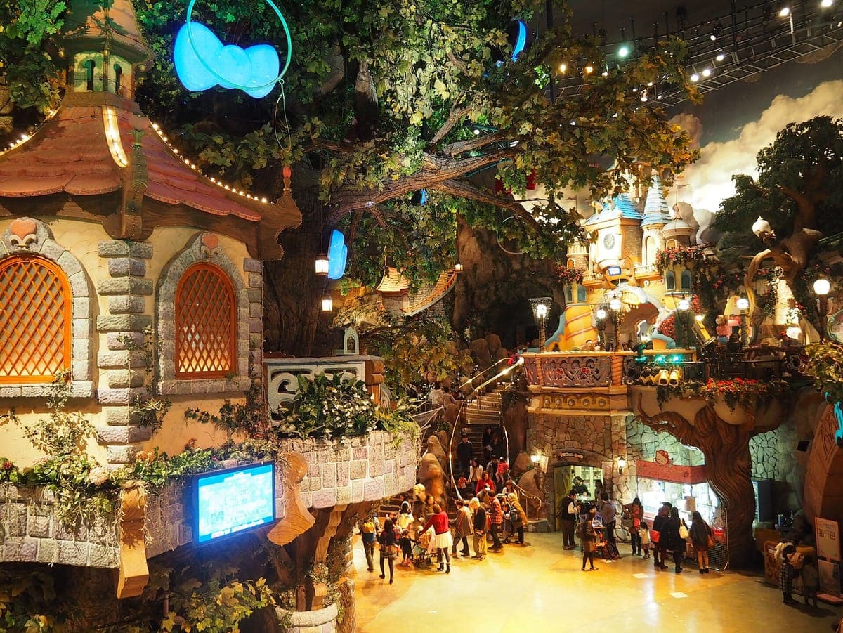 Das Indoor-Puro-Dorf im Sanrio Puroland mit märchenhaften Hausfassaden, einem großen künstlichen Baum mit Lichtern und Besuchern, die auf der Etage darunter laufen