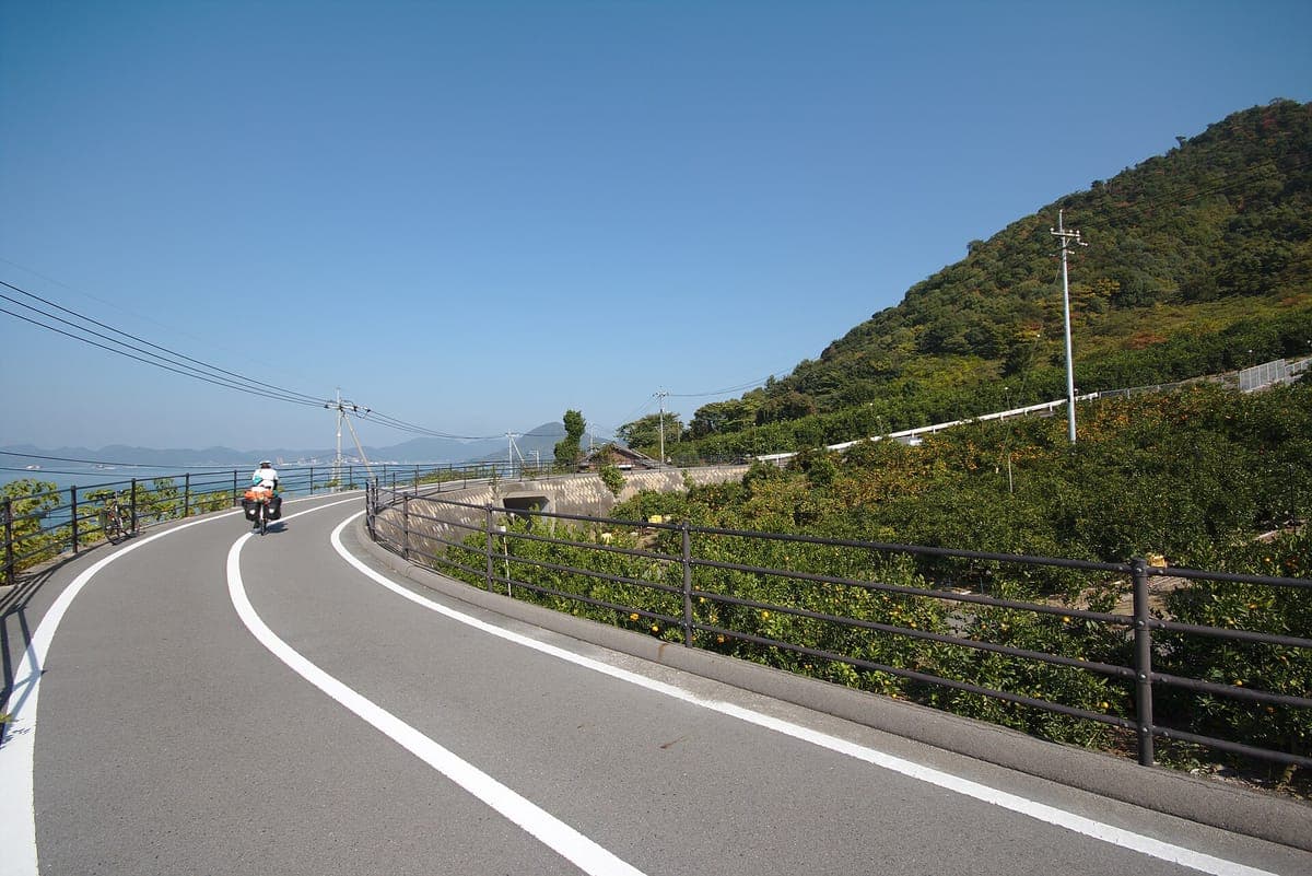 Hladká cyklostezka zakřivená po zeleném svahu nad mořem směrem k vysoké věži lanového mostu s cyklistou v dálce