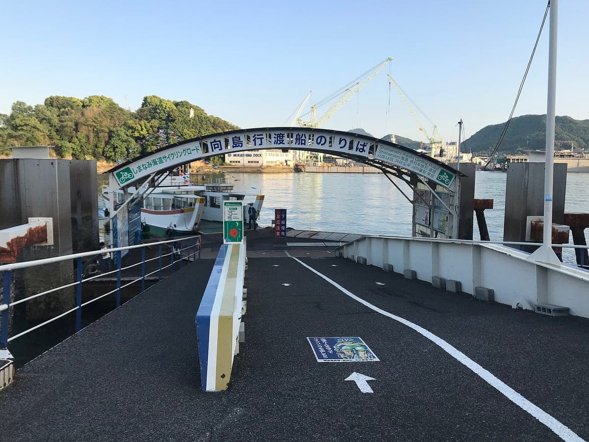 Přívozní molo poblíž nádraží Onomichi s malovaným obloukem přes nástupní rampu, zábradlí a malý trajekt kotvící na konci