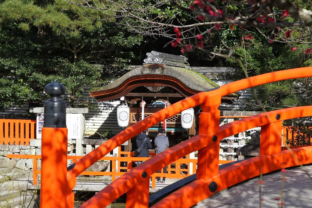 Un pequeño puente con arco bermellón frente a Mitarashi-sha en Shimogamo Jinja, un santuario de madera con linternas blancas, entre árboles.