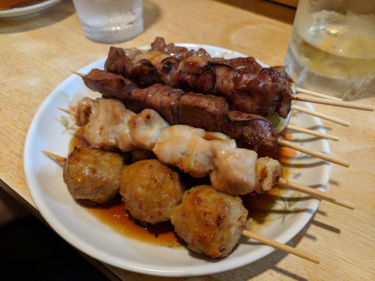 Un plato blanco de brochetas a la parrilla, que incluyen albóndigas, pollo e hígado, sobre una encimera de madera en Omoide Yokochō