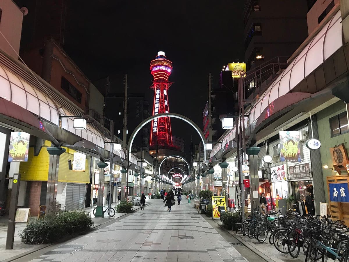 Tsūtenkaku torre iluminada em vermelho à noite no final de uma rua comercial coberta em Shinsekai, emoldurada por um arco de luzes