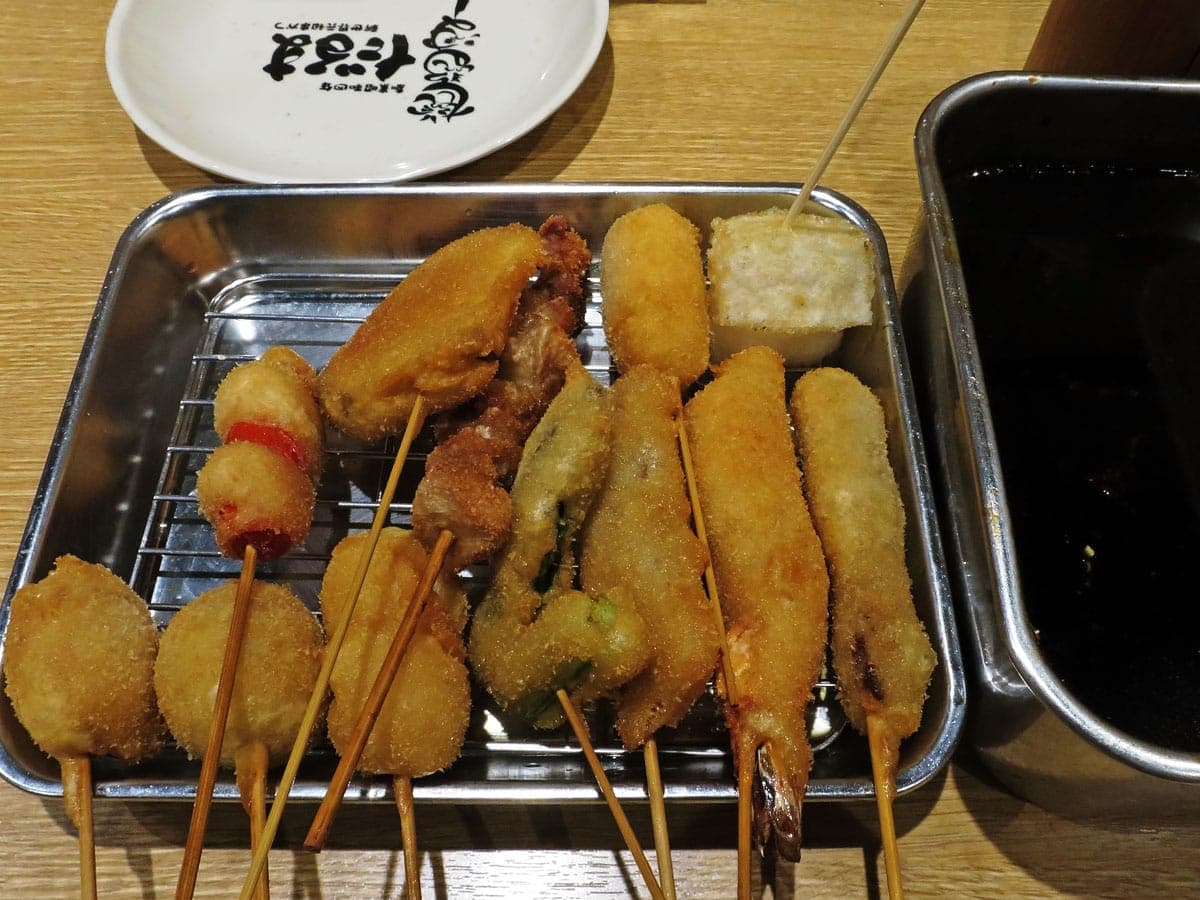 Uma bandeja de metal com espetos dourados fritos kushikatsu, incluindo camarões e vegetais, ao lado de um recipiente de aço com molho