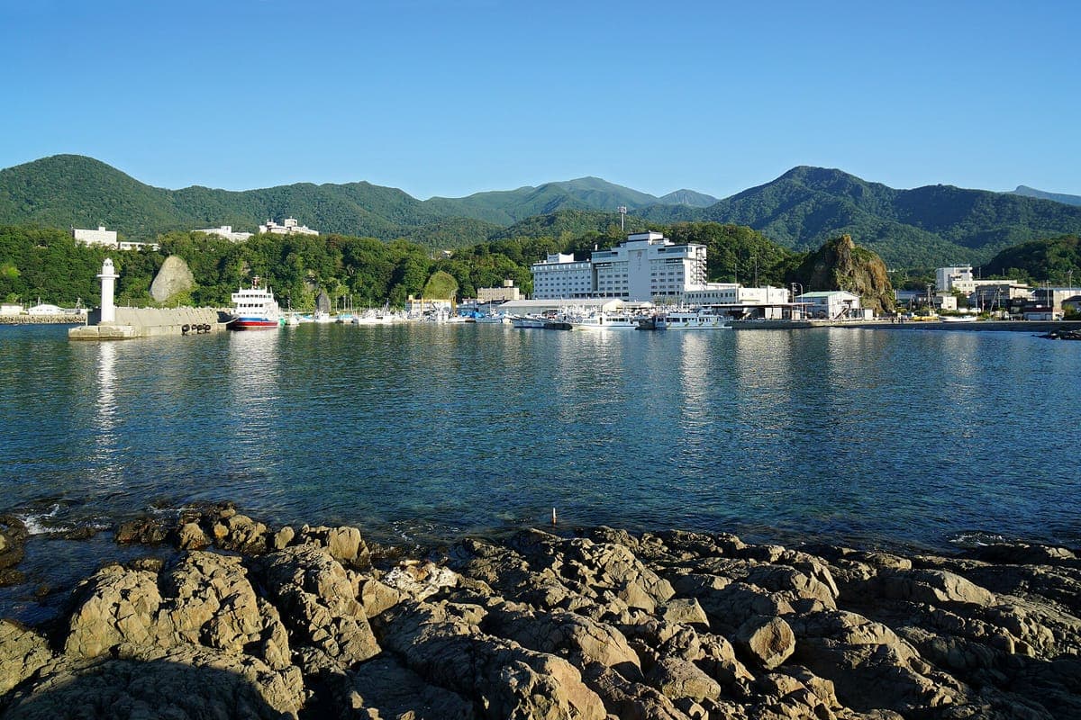 Utoro port sa Shiretoko Peninsula, tahimik na asul na tubig na may mga bato sa harapan, isang maliit na parola, mga bangka at hotel sa ibaba ng mga kagubatan na burol