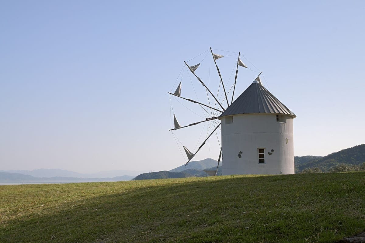 Bílý větrný mlýn v řeckém stylu s trojúhelníkovými plachtami stojící na travnatém svahu v Shōdoshima Olive Park, s mořem a kopci vzadu pod bledou oblohou