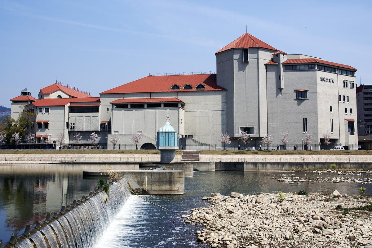 The Takarazuka Grand Theater, a large cream building with red-tiled roofs and towers, seen across a river with a weir, rocks and green hills behind