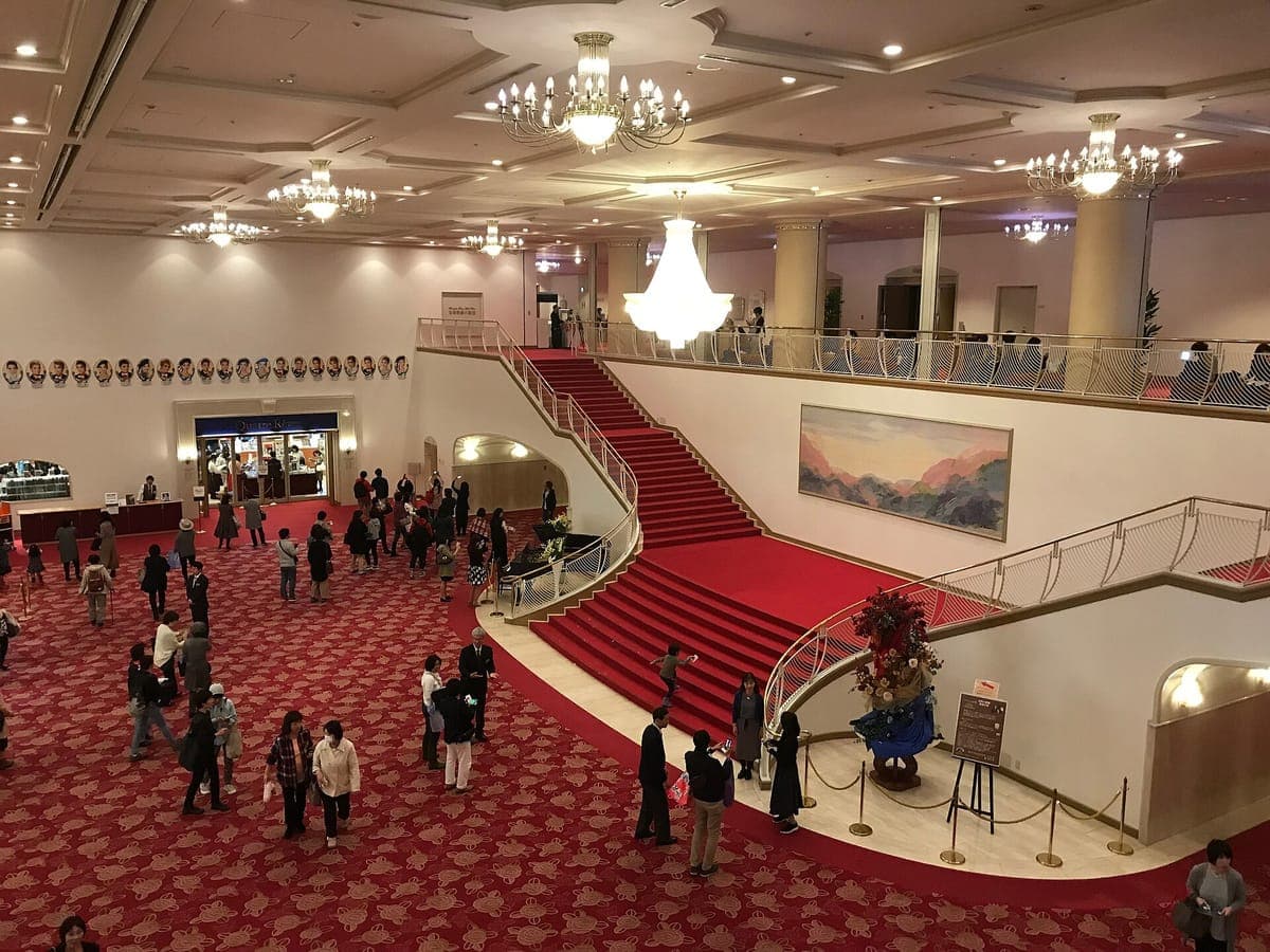 The lobby of the Takarazuka Grand Theater with a curving red-carpeted double staircase, chandeliers, a painting on the wall and visitors on a red patterned carpet