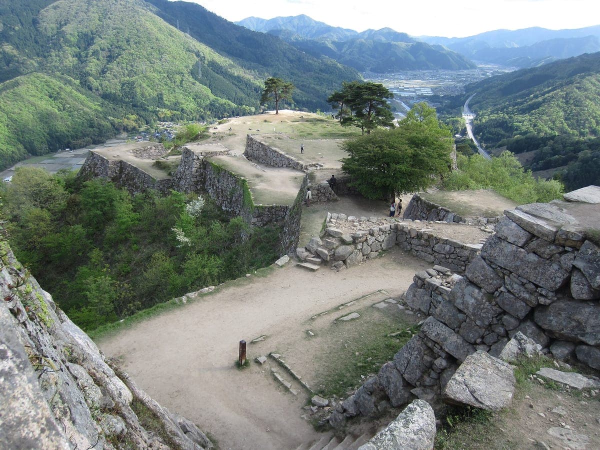Melihat ke bawah dari atas runtuhan Istana Takeda di atas tingkatan dinding batu kelabu dan teres berumput, dengan pokok pain, gunung hijau dan lembah sungai di seberangnya