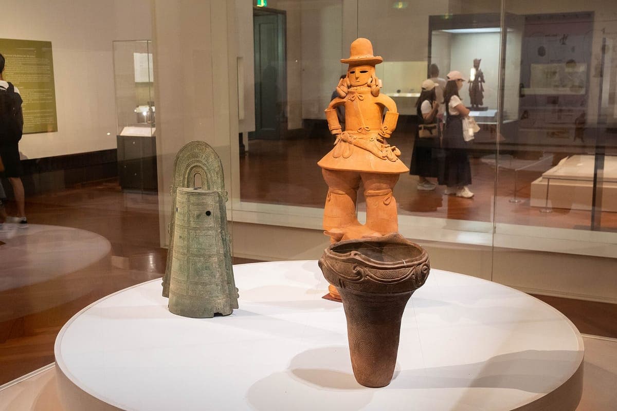 An orange clay haniwa figure of a man in a brimmed hat and a green bronze bell displayed on a white plinth in a museum gallery