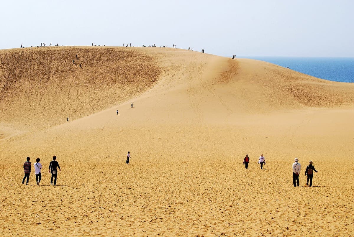 Besökare som går över bred gyllene sand mot den långa sluttningen av en stor sanddyn vid Tottori, med små figurer längs åsen och en remsa av blått hav till höger