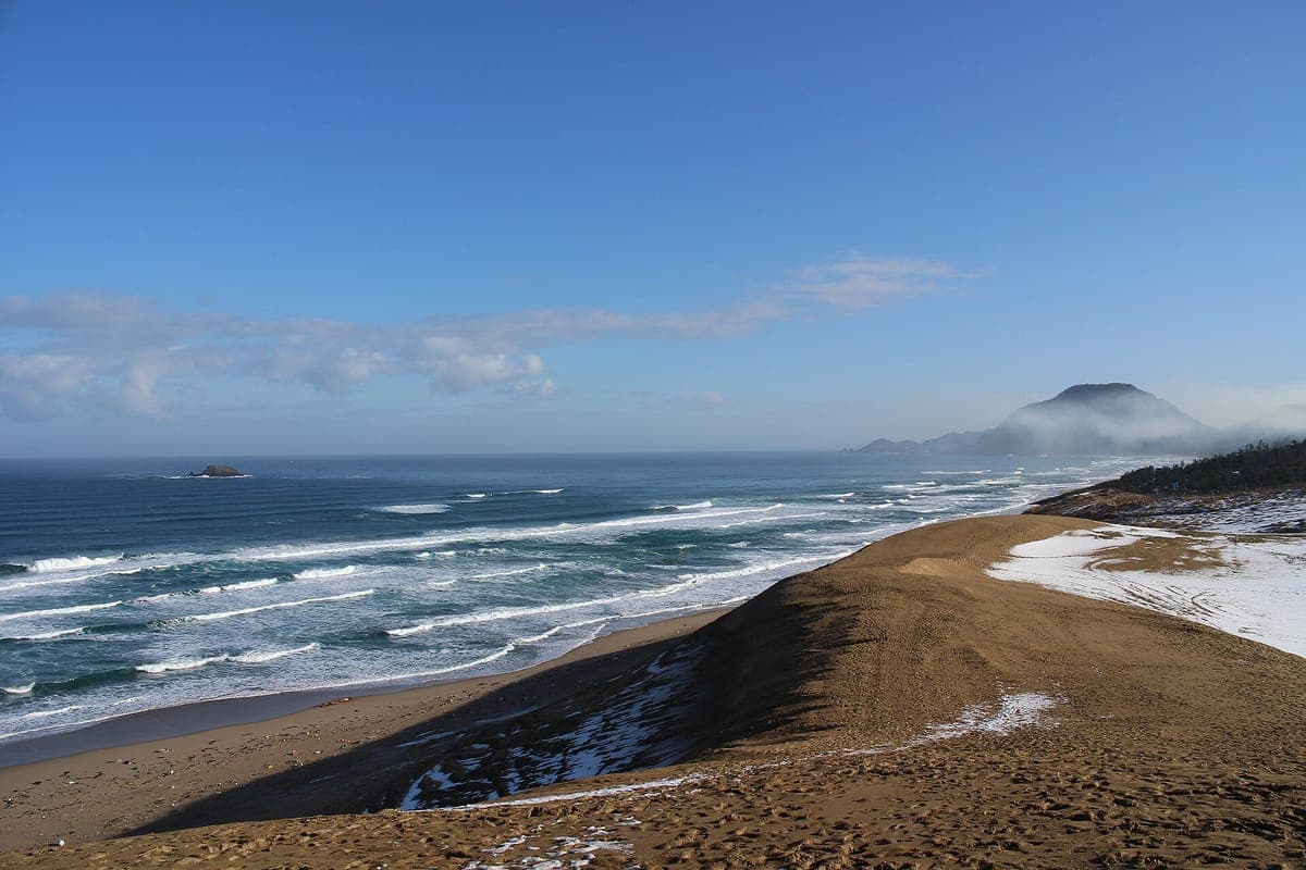 Krönet av en Tottori-sanddyn dammad av snö, sluttande ner mot en strand med vittäckta vågor under en klarblå himmel, med en udde vid horisonten