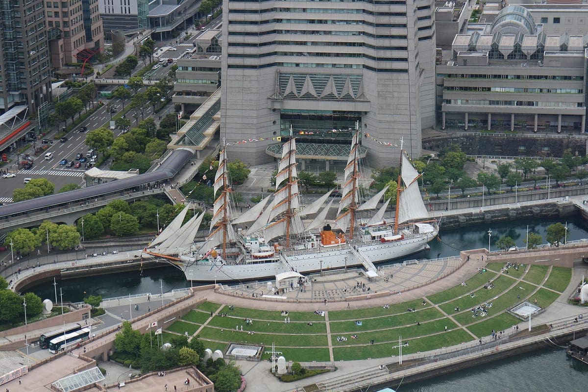Vista aérea del velero blanco de cuatro mástiles Nippon Maru en su muelle de piedra junto a un césped curvo, con el complejo Landmark Tower detrás