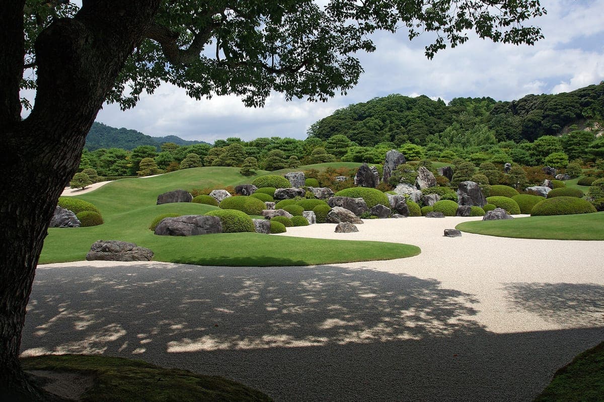 Der trockene Landschaftsgarten des Adachi Museum of Art mit geharktem weißem Kies, runden moosigen Hügeln, Felsen und gestutzten Kiefern, dahinter bewaldete Hügel, eingerahmt von einem dunklen Baumstamm und Ästen