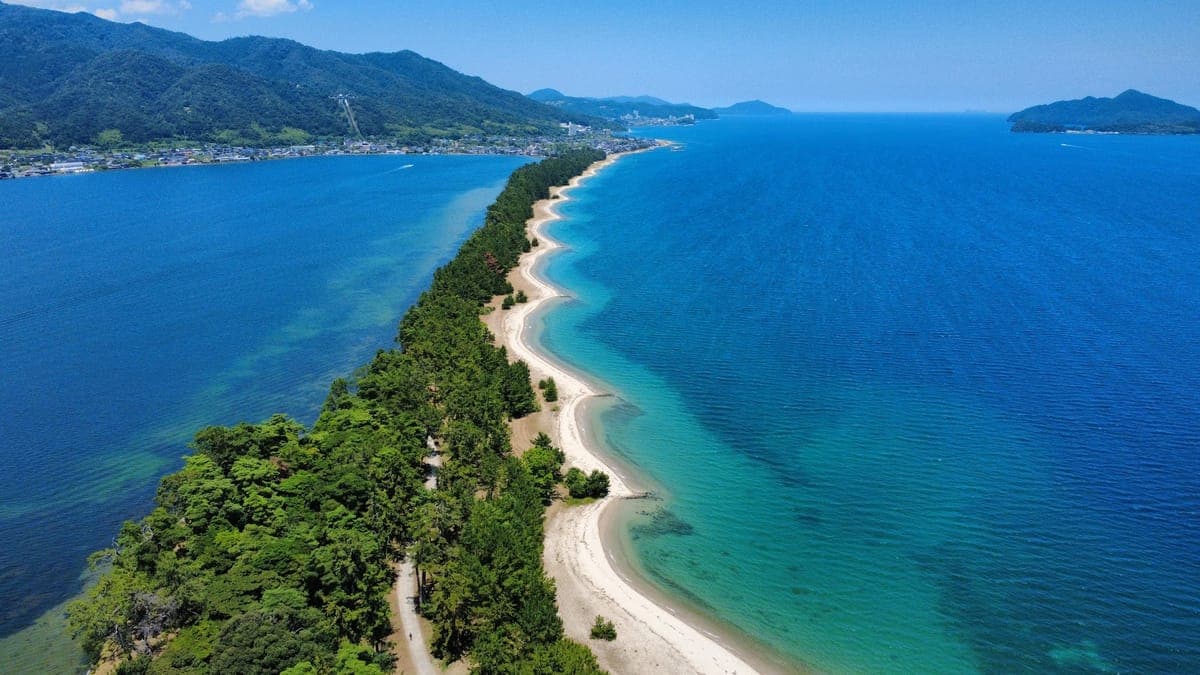 Aerial view along the narrow Amanohashidate sandbar, with a path through dense pines, a white sand beach and turquoise water on one side