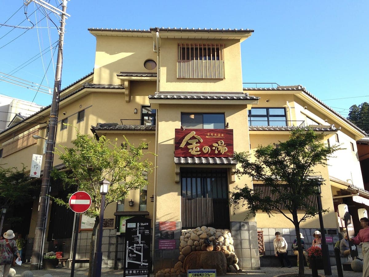 Les bains publics Kin no Yu d'Arima Onsen, un bâtiment de couleur crème avec un toit de tuiles traditionnel et un panneau rouge indiquant 金の湯, dans une rue ensoleillée