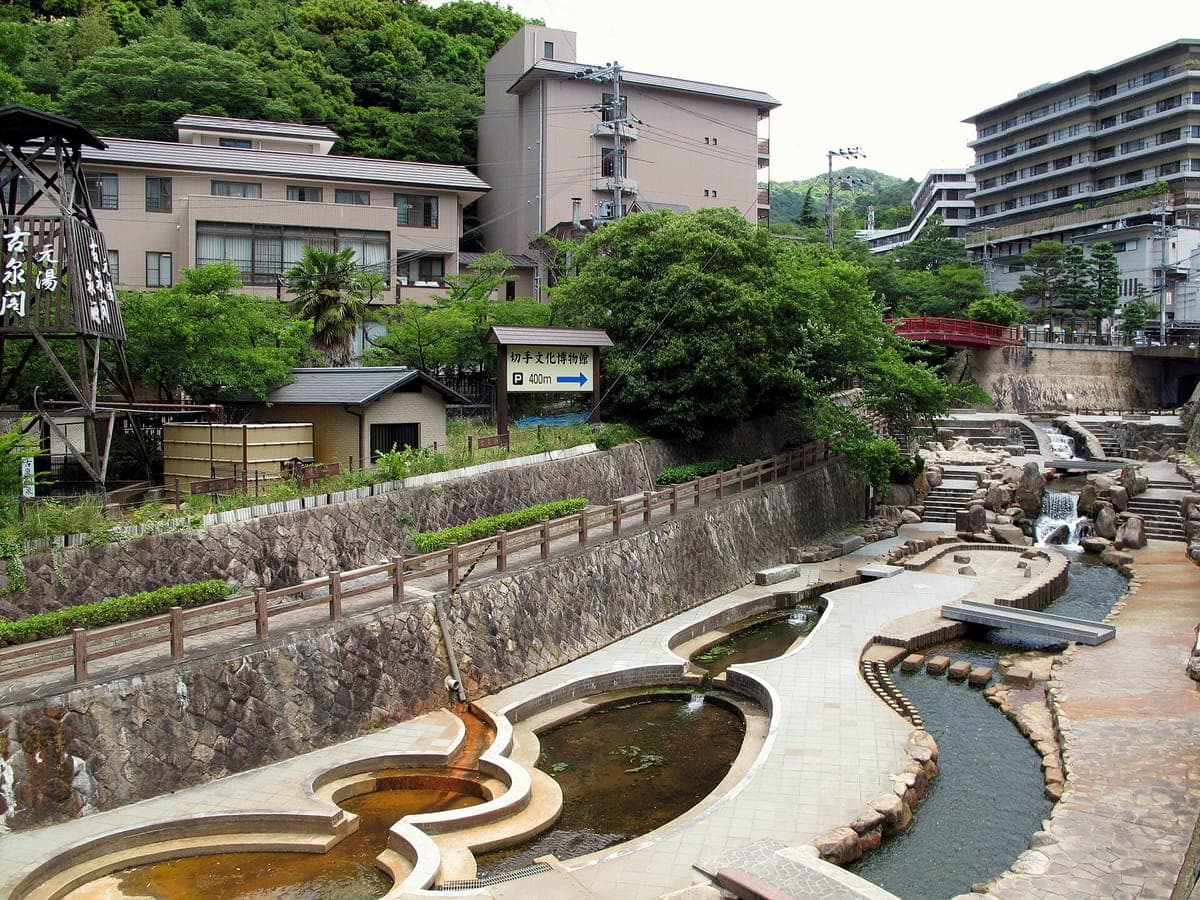 La rivière Arima bordée de pierres coule dans des canaux incurvés au milieu d'Arima Onsen, avec un petit pont rouge et des hôtels sur la colline verdoyante derrière