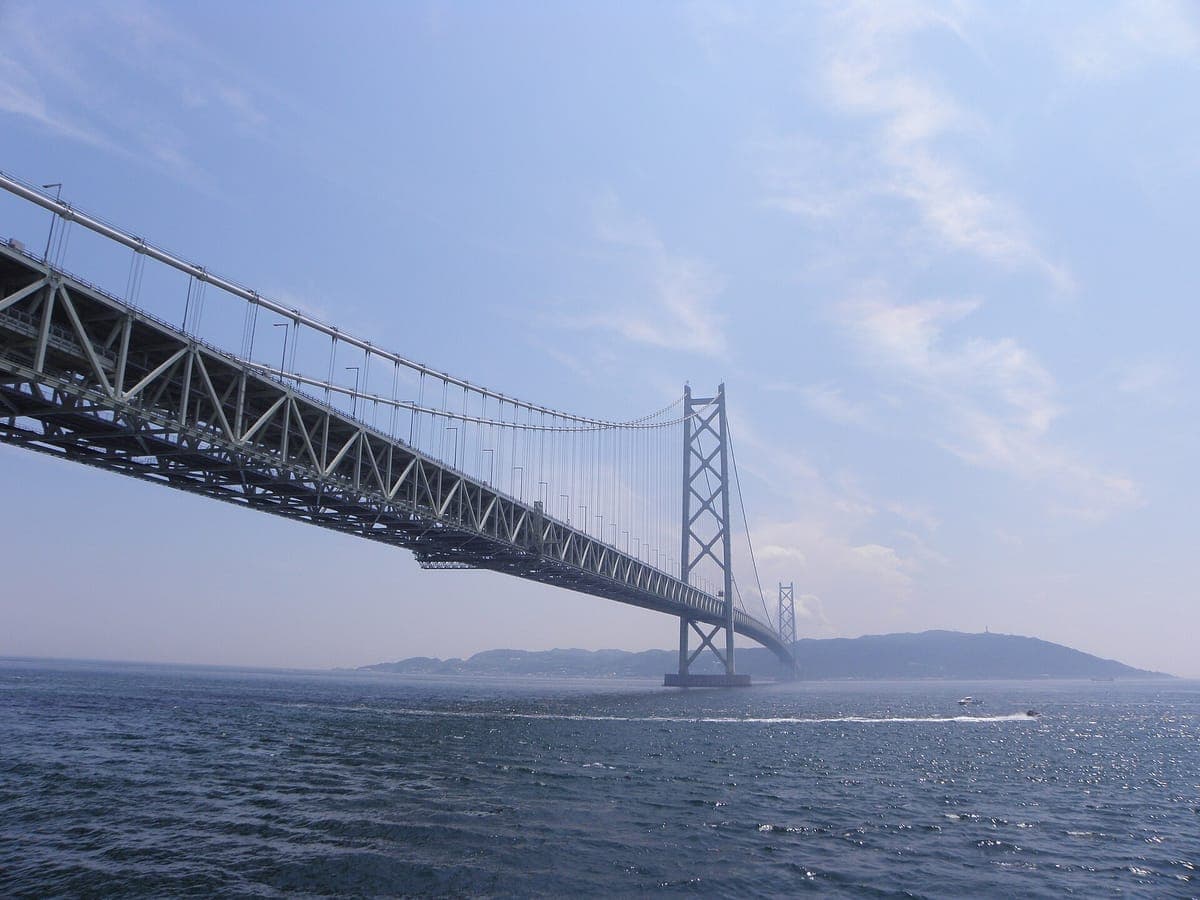 Die Akashi-Brücke Kaikyō, die sich von der Akashi-Seite aus über die Meerenge erstreckt, mit einem hohen weißen Turm und Tragseilen über graublauem Wasser und der Insel Awaji im Dunst