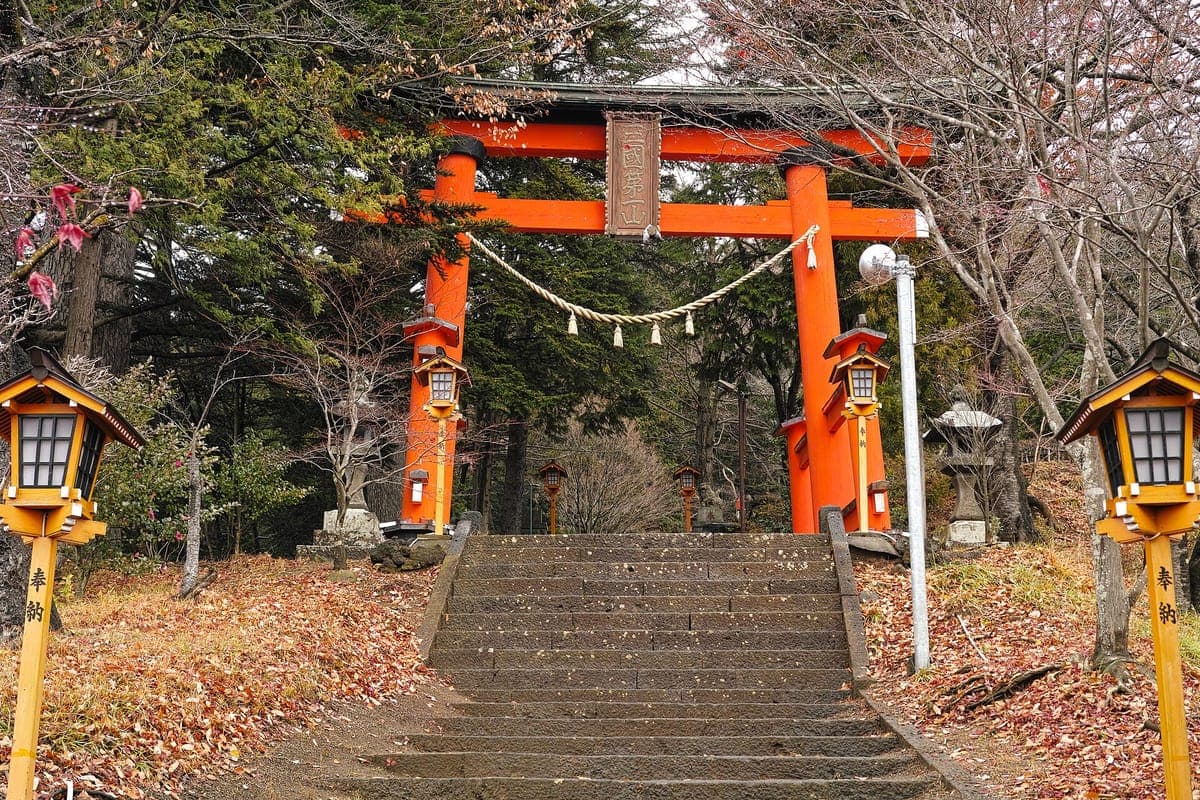 Světlá rumělková brána torii na vrcholu kamenných schodů ve svatyni Arakura Fuji Sengen, lemovaná žlutými lucernami, s holými stromy a spadaným listím
