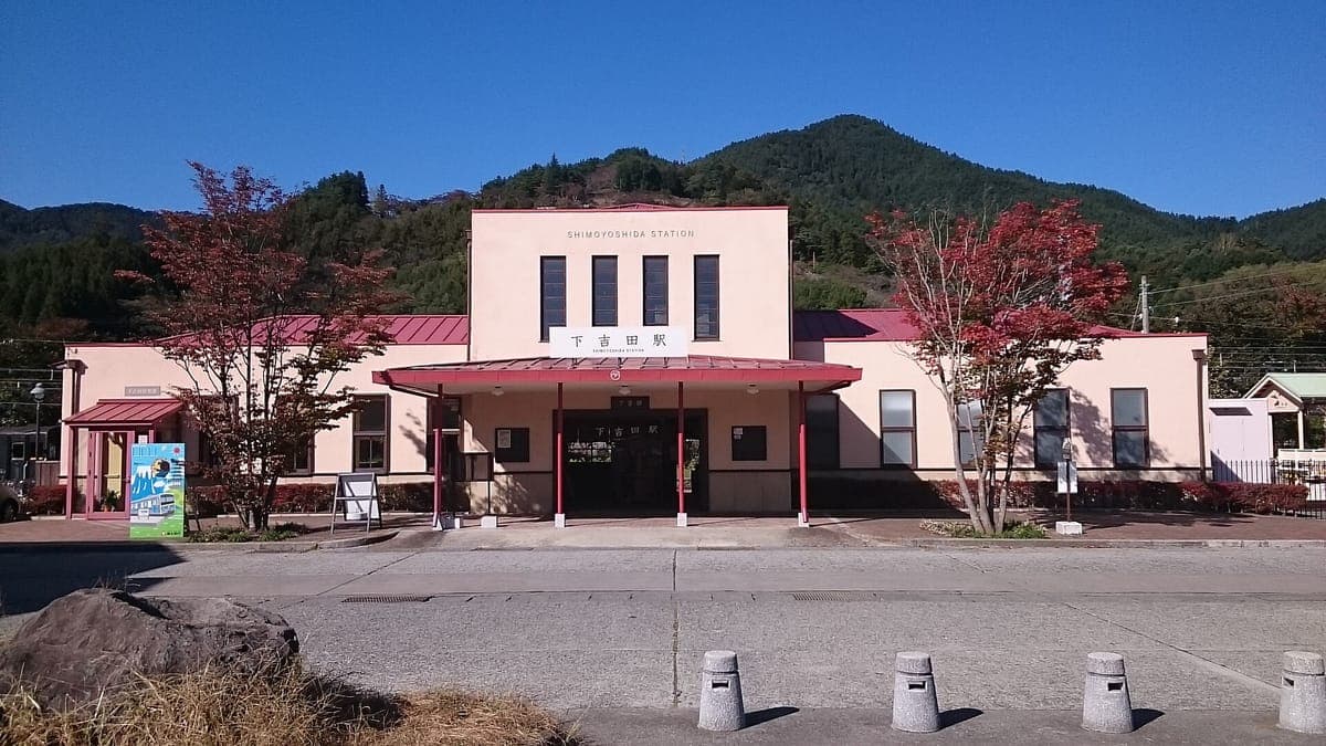 Malá růžová a bílá budova stanice Shimoyoshida na lince Fujikyu pod jasně modrou oblohou s podzimními stromy a zelenými kopci za