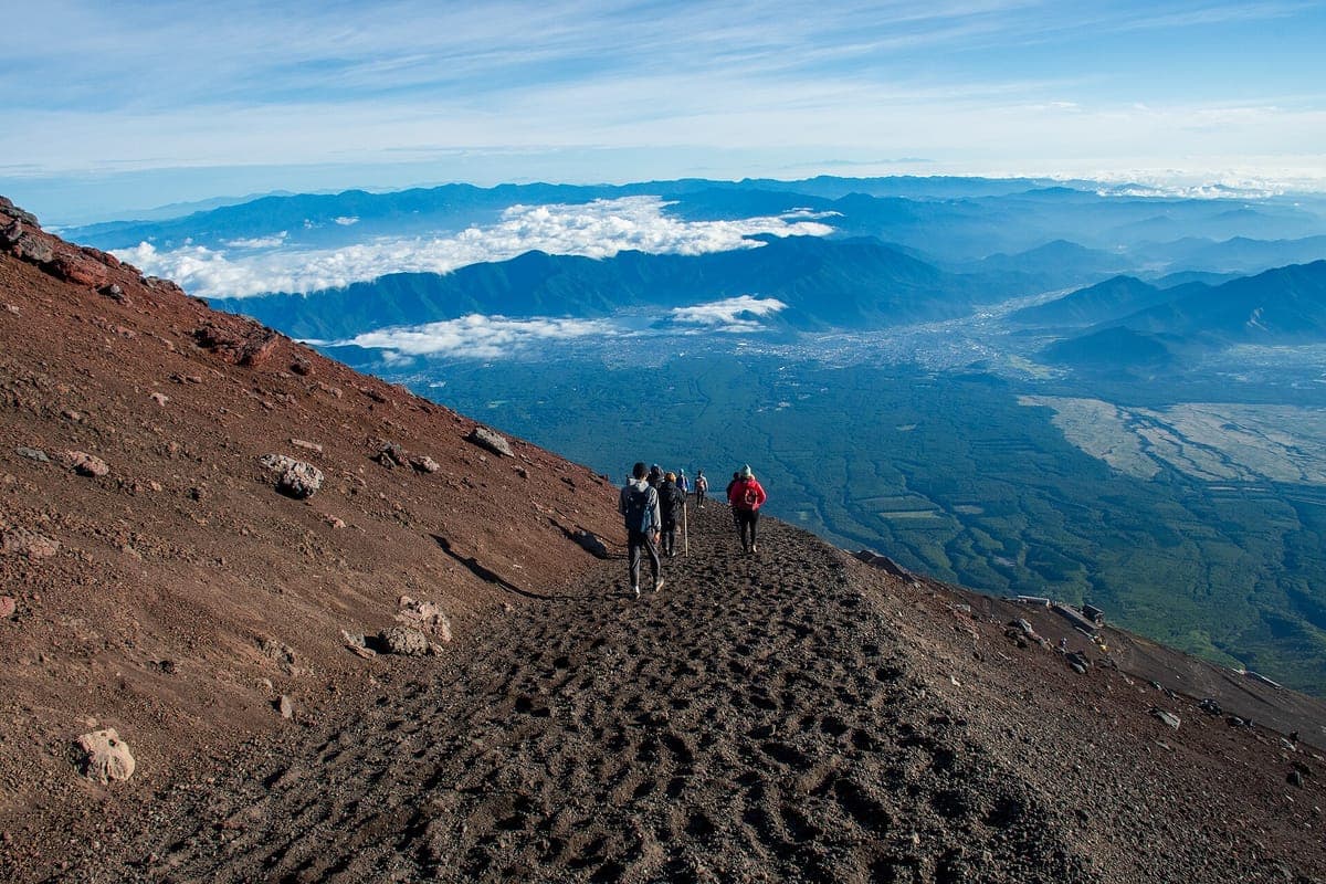Wandelaars lopen een kale roodbruine vulkanische helling op op de Yoshida Trail van de berg Fuji, met daaronder een zee van wolken en verre bergen