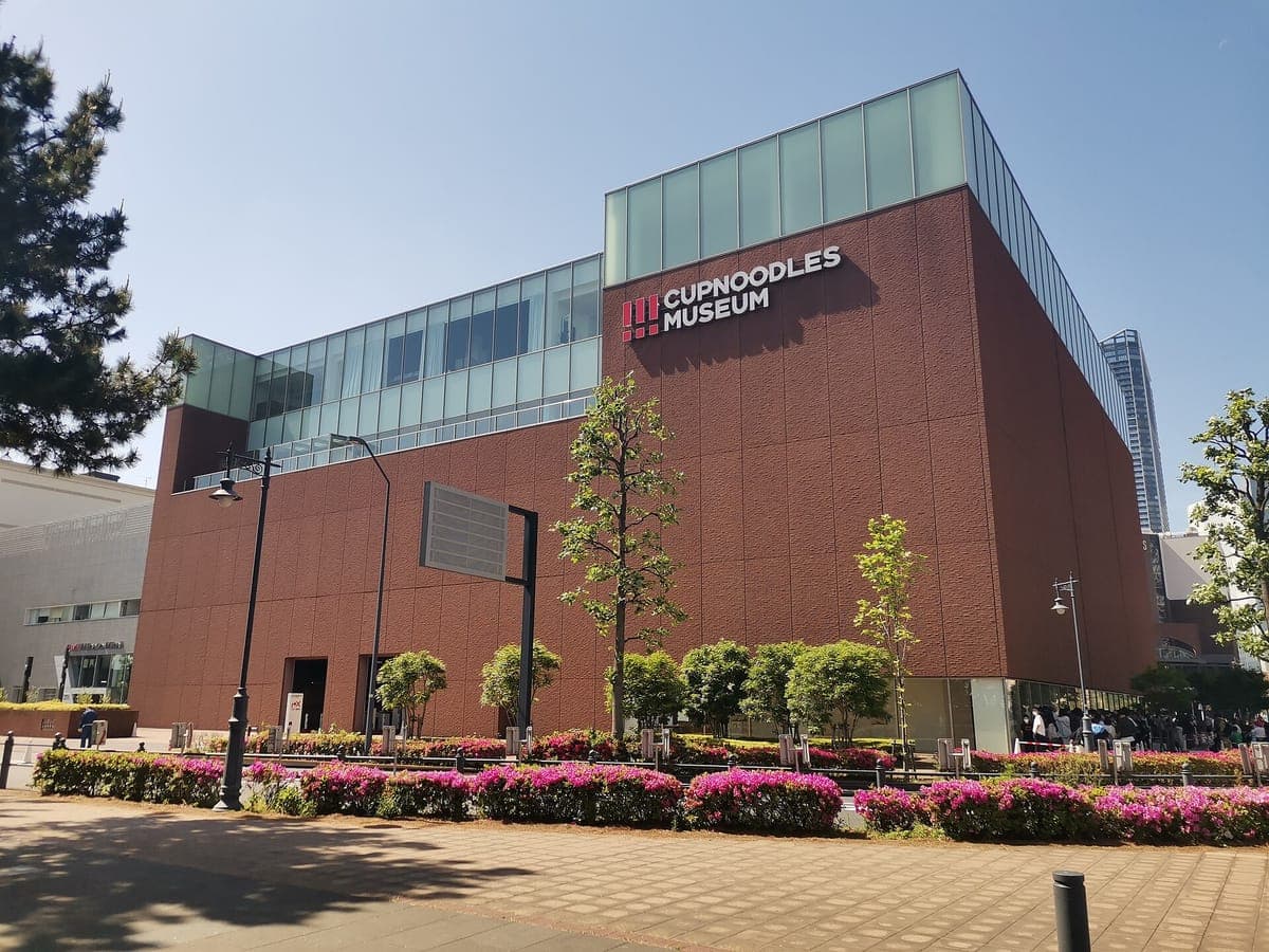 Das ziegelrote, kastenförmige Cup Noodles Museum in Yokohama mit einem Glasgeschoss und dem Namen des Museums an der Wand, dahinter Bäume und rosa blühende Sträucher
