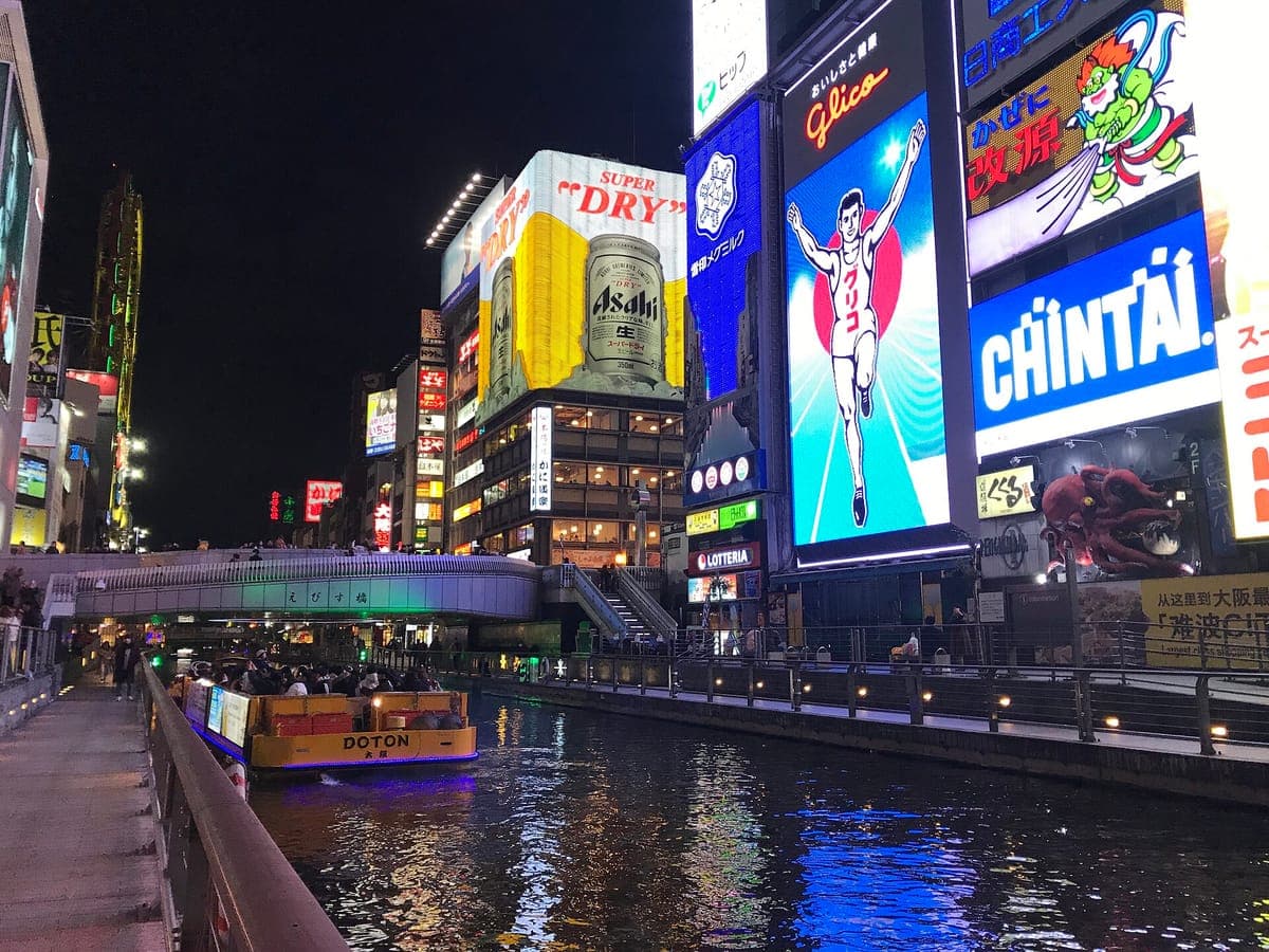 Ang maliwanag na Glico running man sign at iba pang neon billboard sa itaas ng Dōtonbori canal sa gabi, na may sightseeing boat sa tubig