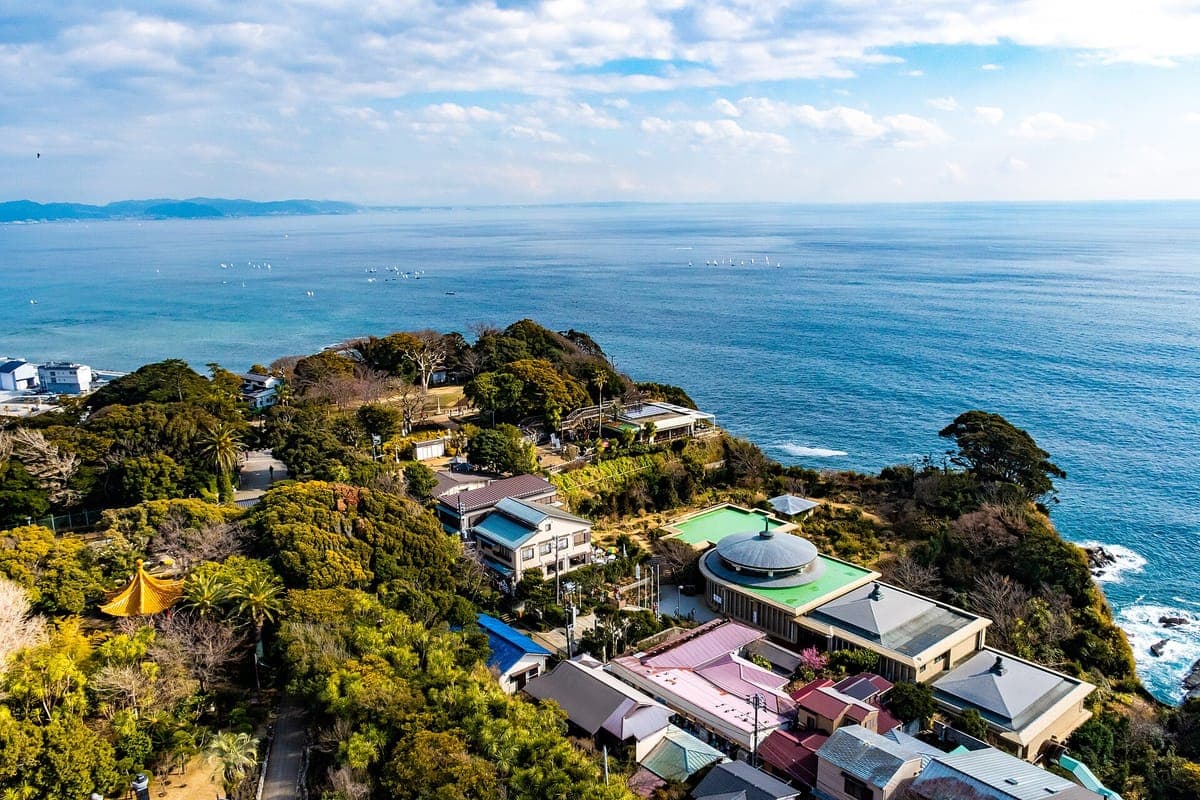 Pohled z Enoshima Sea Candle přes zalesněný vrchol kopce Enoshima, jeho střechy a zahrady a modré moře táhnoucí se ke vzdálenému pobřeží