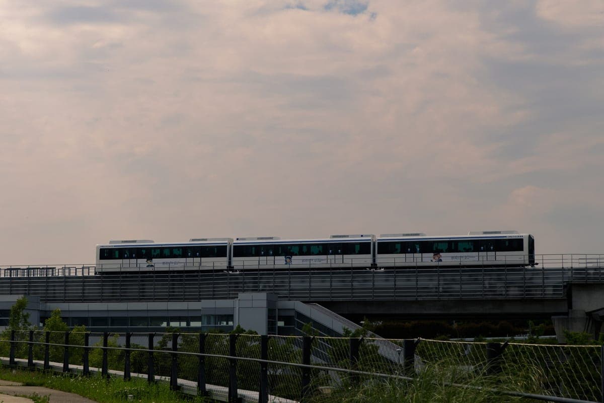 Isang puting tatlong-kotse na Linimo maglev na tren na tumatakbo sa isang mataas na riles sa ilalim ng maulap na kalangitan