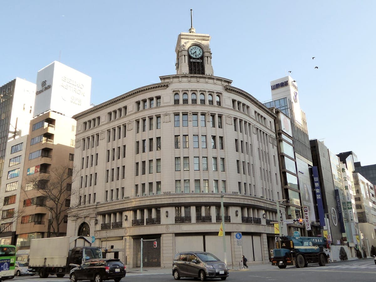 Zakřivená kamenná budova Wakō s hodinovou věží na rohu křižovatky Ginza 4-chōme s moderními budovami a auty kolem ní