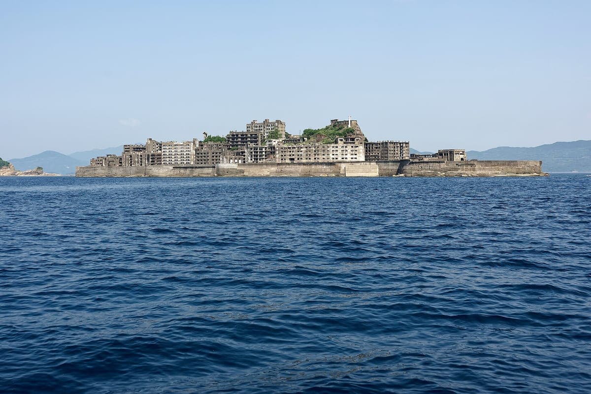 Hashima denizden görülen ada, deniz duvarının arkasında, mavi gökyüzünün altında, yıkık beton binalarla kaplı uzun alçak bir ada