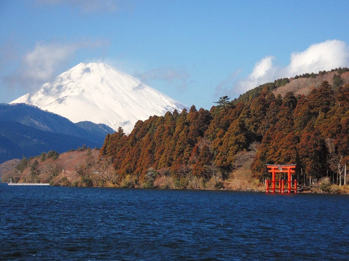 Zasněžená hora Fuji tyčící se za zalesněným svahem na jezeře Ashi v Hakone s červenou torii stojící na okraji vody