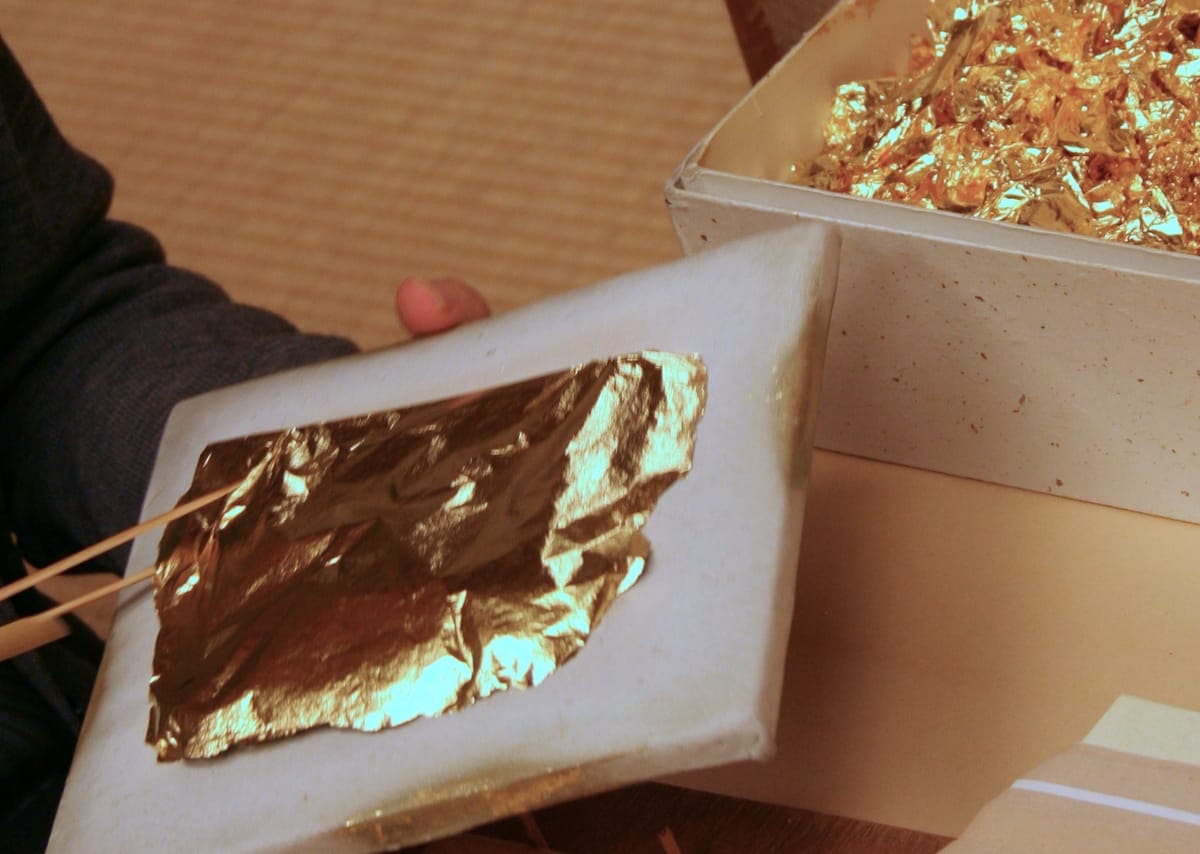 Eine Hand hält ein Blatt Papier mit zerknittertem Blattgold neben einer offenen Schachtel mit Blattgoldflocken