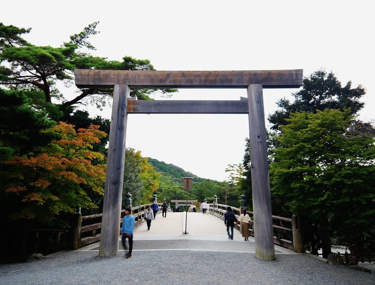 Un torii alto y sencillo de madera en la entrada del Uji-bashi Santuario Interior de Ise Jingū's, con un largo puente de madera y colinas boscosas más allá