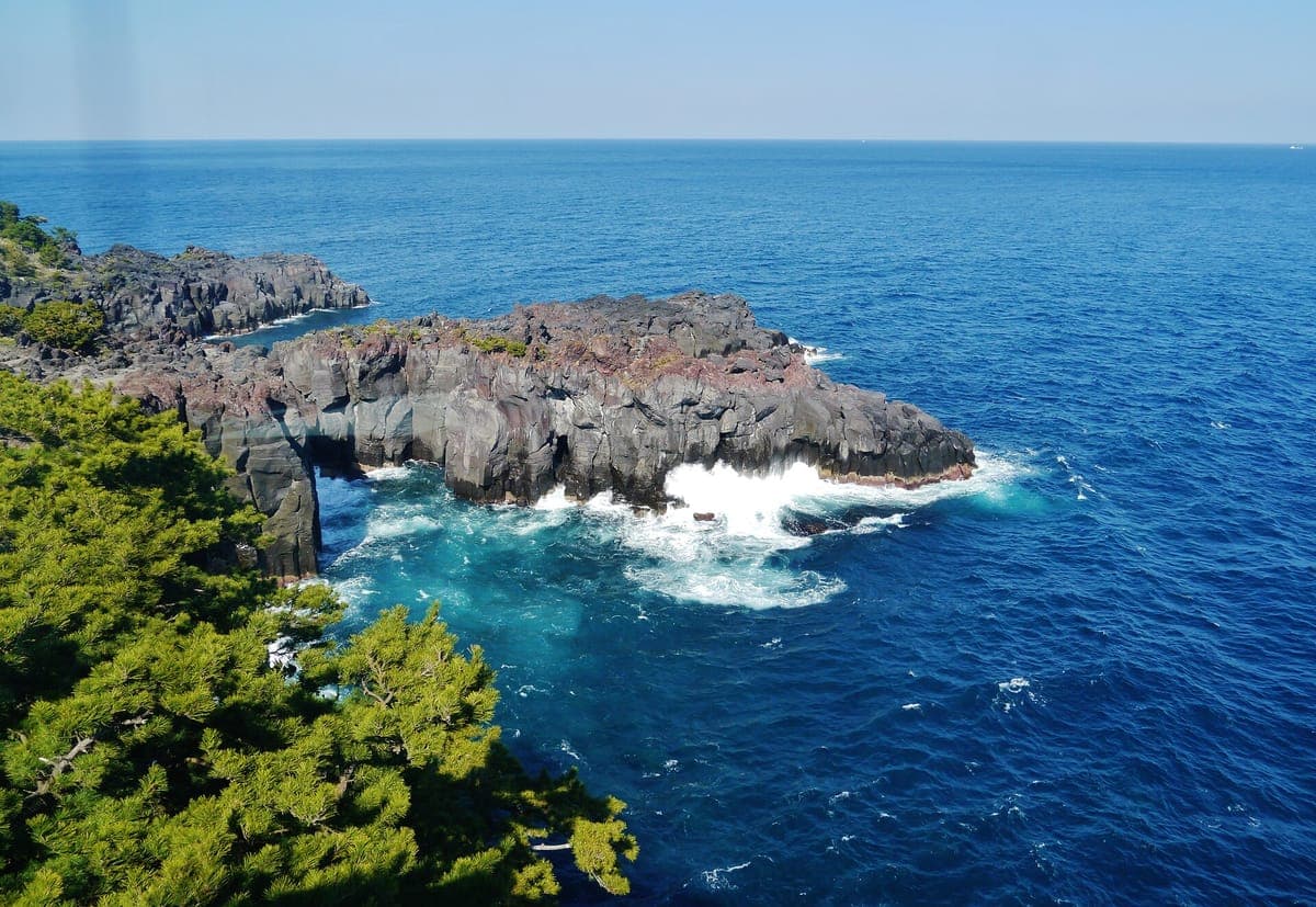 Mga itim na bulkan na batong cliff at isang mabatong headland sa Jōgasaki baybayin, na may mga alon na humahampas sa malalim na asul na dagat at mga pine tree sa harapan