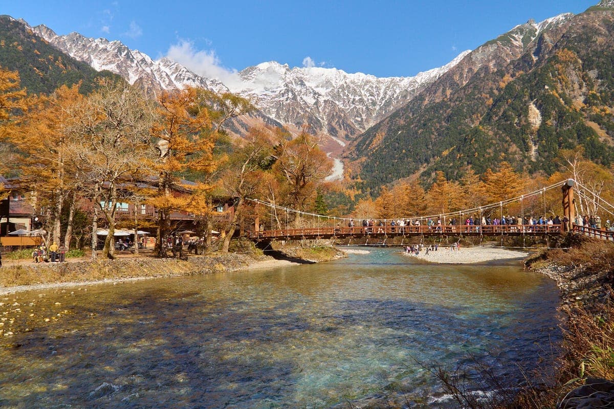 Arkasında turuncu sonbahar karaçamları ve karla kaplı sivri uçlu zirvelerle, berrak Azusa Nehri'ni Kamikōchi'da geçen ahşap Kappa Köprüsü