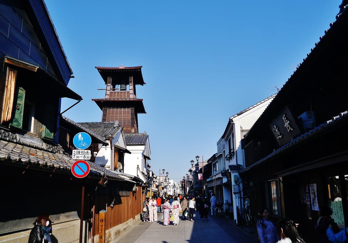 Isang makipot na kalye sa Kawagoe na may linya na may madilim na tiled na mga merchant house, patungo sa matataas na kahoy na Toki no Kane bell tower sa ilalim ng asul na kalangitan