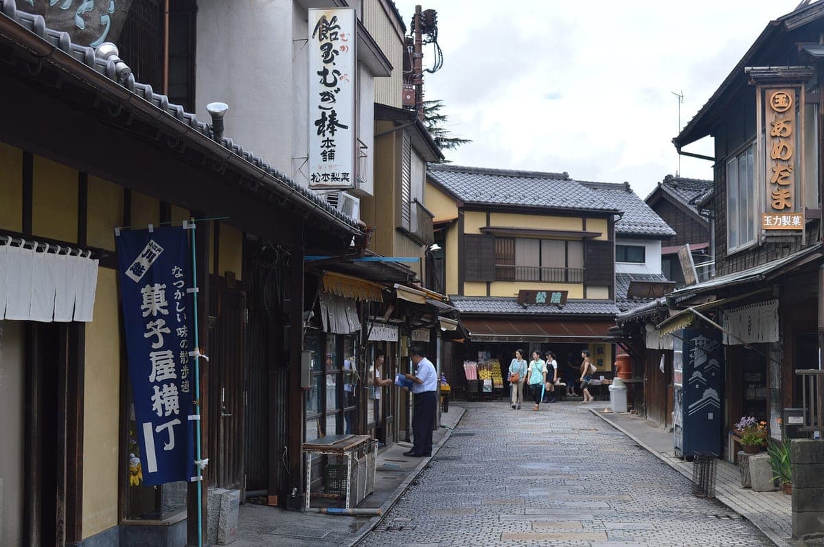 Isang makipot na daang sementadong bato sa Kashiya Yokochō ng Kawagoe na may linya na may mababang kahoy na tindahan ng matamis, nakasabit na mga karatula sa tindahan at mga banner
