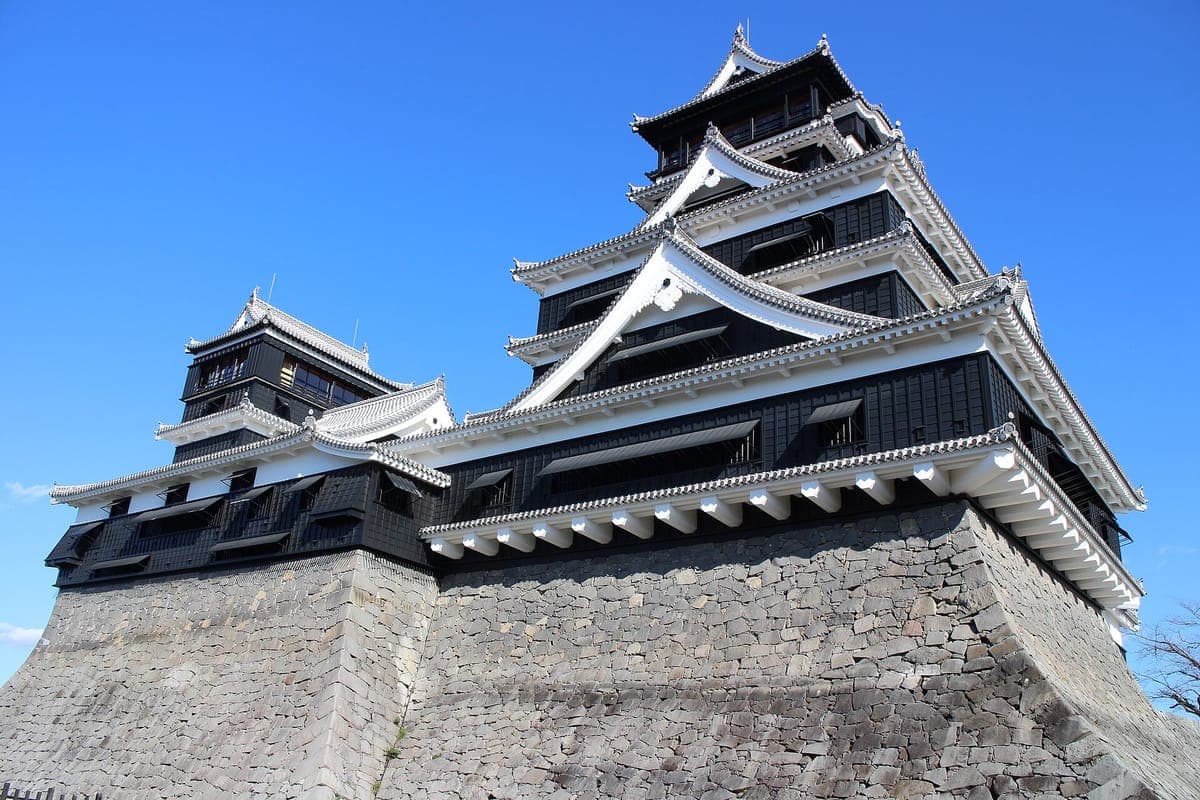 Blízký pohled na hrad Kumamoto zespodu s černými dřevěnými stěnami, bíle lemovanými střechami a vysokou zakřivenou kamennou základnou