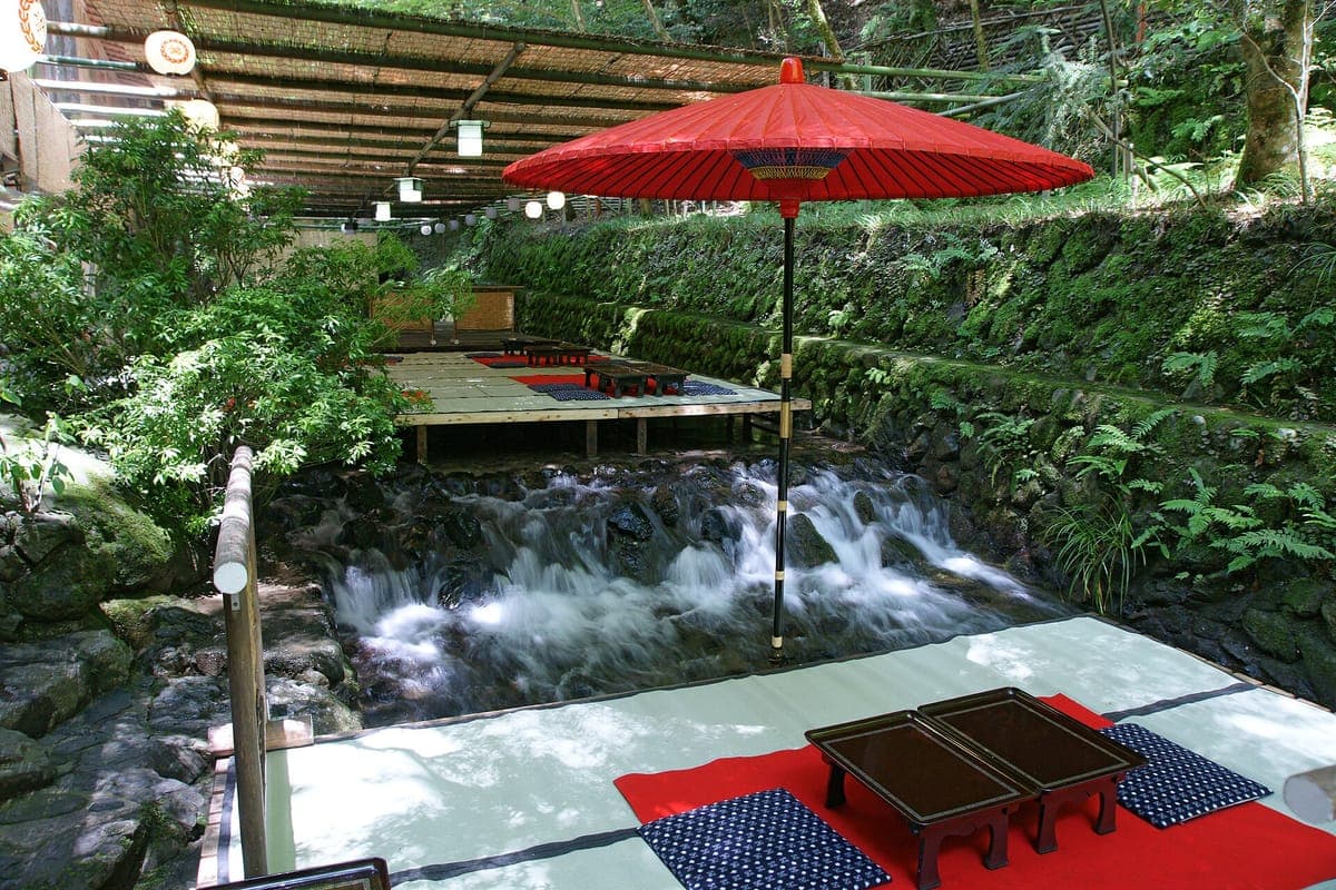 Uma plataforma de jantar de tatame com tapetes vermelhos e mesas baixas construída sobre uma pequena cachoeira no riacho Kibune, à sombra de um guarda-sol vermelho