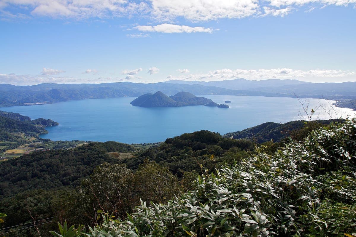 See Tōya von einem Hügel aus gesehen, ein breiter blauer See mit einer Gruppe bewaldeter Inseln in der Mitte, grünen Hügeln im Vordergrund und Bergen am anderen Ufer