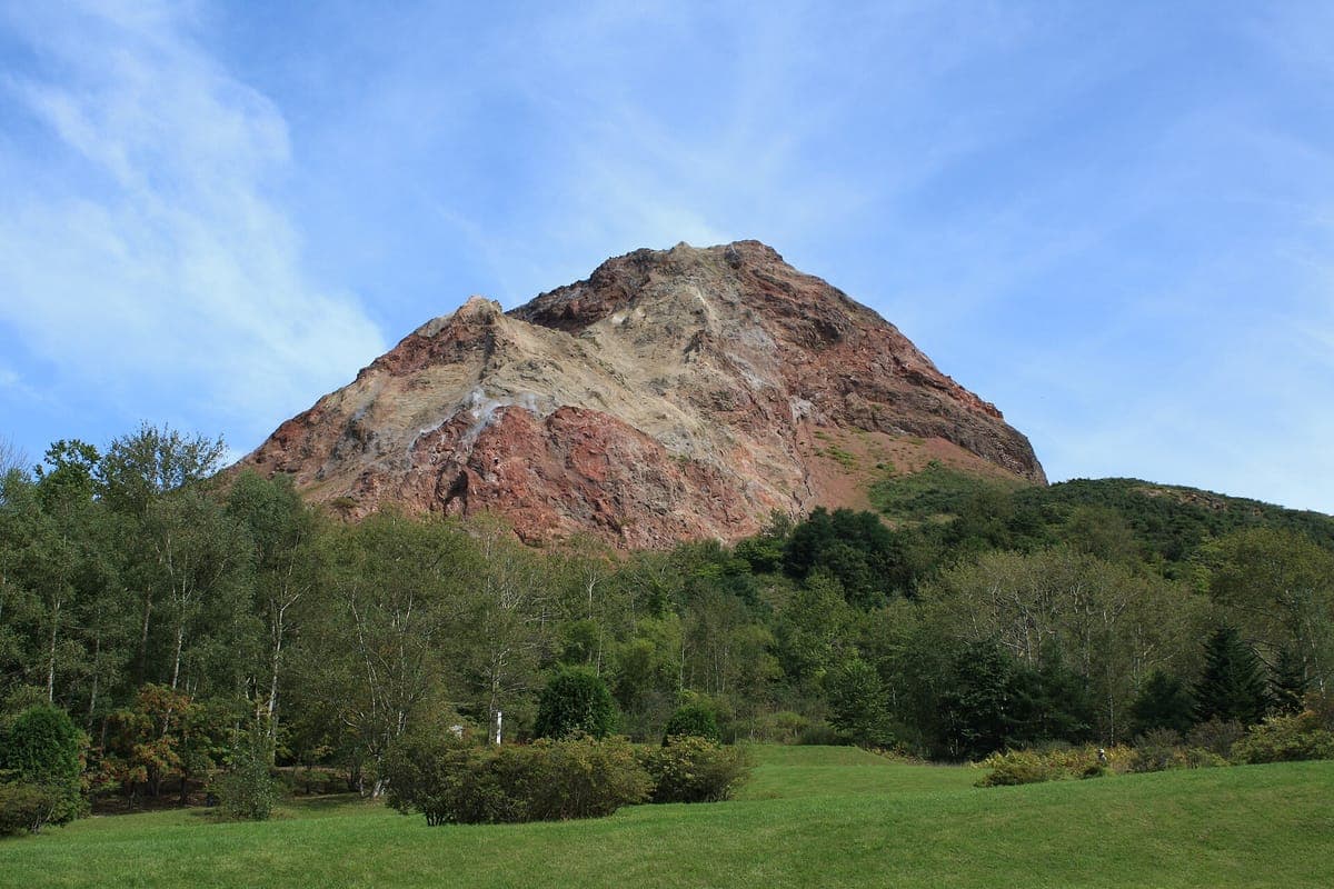 Shōwa-shinzan, ein abgerundeter rotbrauner Vulkangipfel mit kahlen Felshängen, die sich über grünen Bäumen und einer Wiese unter blauem Himmel erheben
