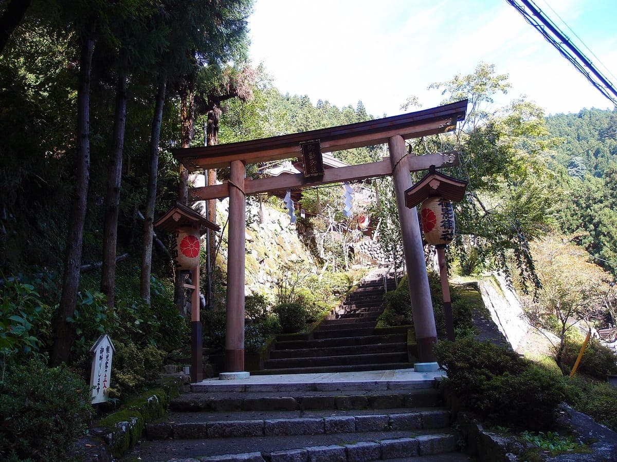 Ein zinnoberrotes Torii, flankiert von zwei Laternen, am Fuße einer Steintreppe, die in den Wald auf dem Mount Mitake führt, mit einem weißen Wegweiser auf der linken Seite