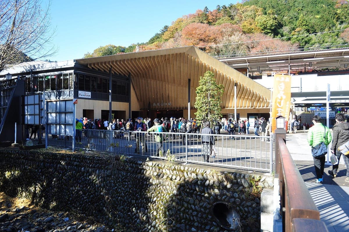 Takaosanguchi Nádraží s velkou šikmou dřevěnou střechou nad vchodem, dav turistů vpředu a podzimně zbarvené kopce za nimi